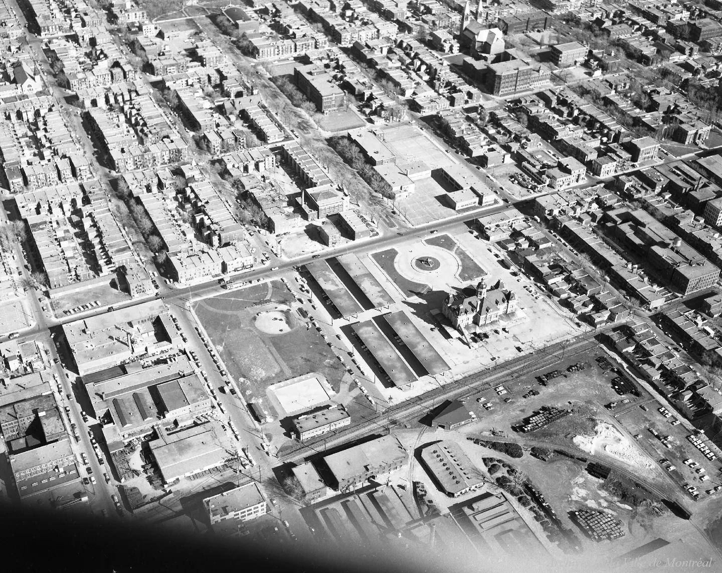 Photo en noir et blanc d’une rue de Montréal, bâtiments alignés, parc au centre, piétons et voitures sur le trottoir.