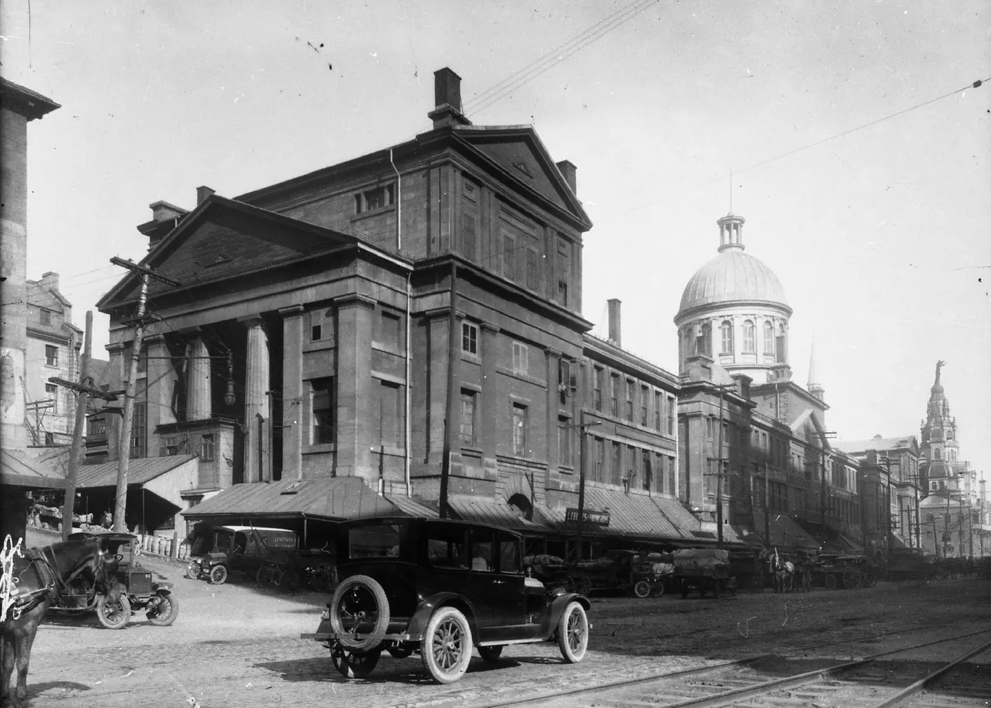 Photo en noir et blanc d’une rue de Montréal avec un bâtiment à tour de l’horloge, des voitures anciennes et des passants.