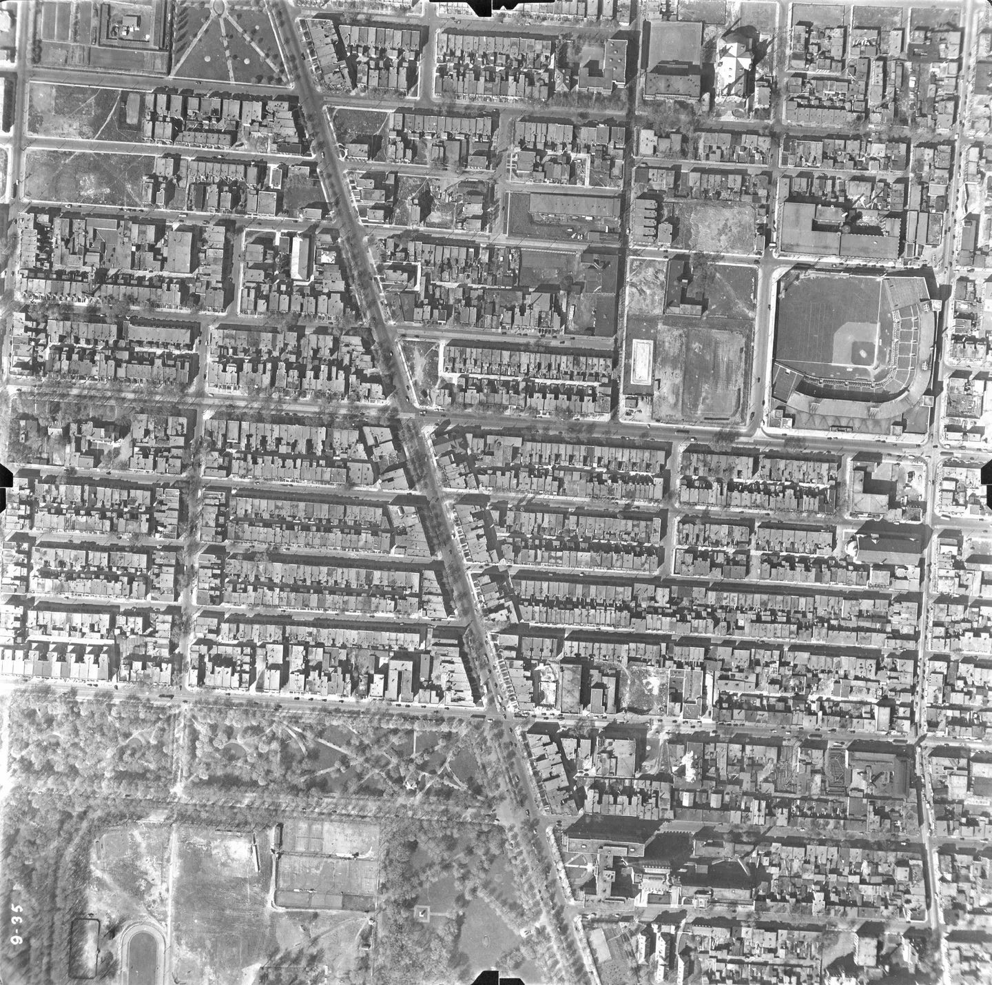 Aerial view of a street with buildings, parked cars and a baseball field in the background